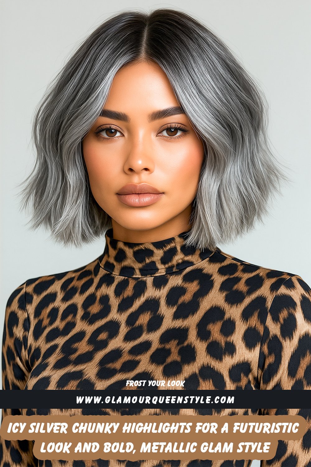 Woman with medium-length dark hair styled in chunky icy silver highlights over smoky roots; wearing sculpted makeup focused on highlighted skin and graphic liner; dressed in a metallic silver sleeveless gown with deep V neckline.